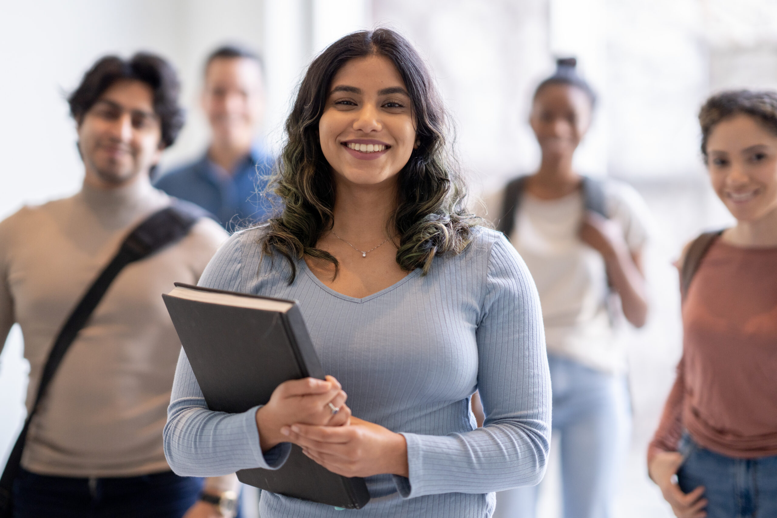 A small group of five University students stop in the hallway of the school to pose for a portrait.  They are each spread out evenly and dressed casually with backpacks and book bags over their shoulders, as they smile for the portrait.