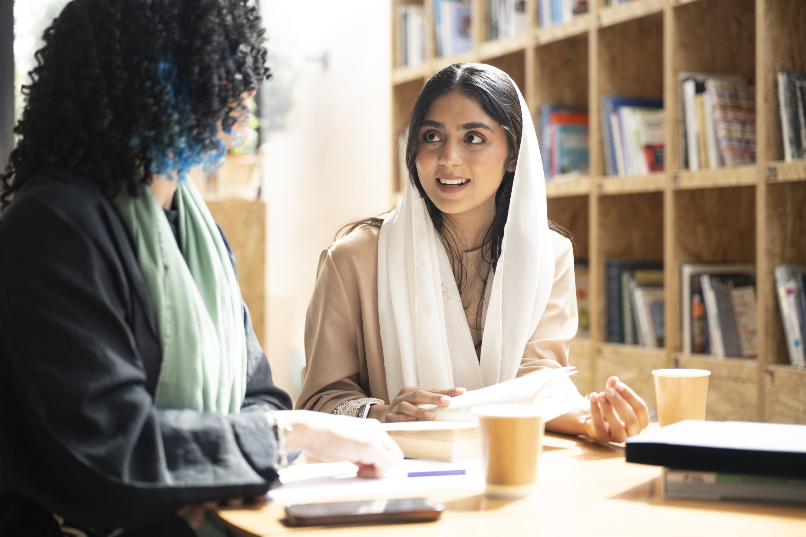 Traditionally dressed students sitting at table with research books, notebooks, and coffee, pausing from working to interact.