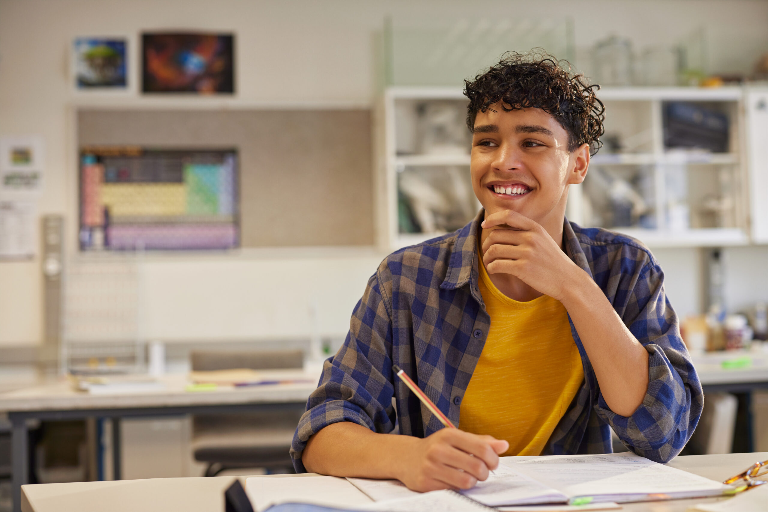 Male teen student smiling while holding a pencil and thinking in class with copy space. Casual teenager student sitting at desk, studying and feeling confident in classroom. Proud and satisfied teen in high school class looking away during lesson with hand on chin.